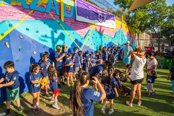 Fotografía de un muro de Boulder de colores azul rosa y amarillo y niños frente a el al aire libre sobre el pasto.