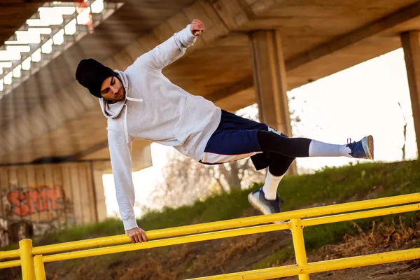 Fotografía de un hombre con sudadera blanca y gorro negro practicando parkour en un barandal color amarillo.