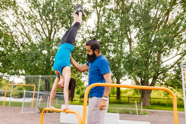 Mujer parada sobre sus manos en una barra amarilla con ayuda de un profesional durante entrenamiento de parkour.