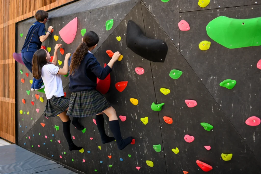 Fotografía de niños con uniforme escolar escalando para practicar Boulder en un muro con agarraderas de colores.